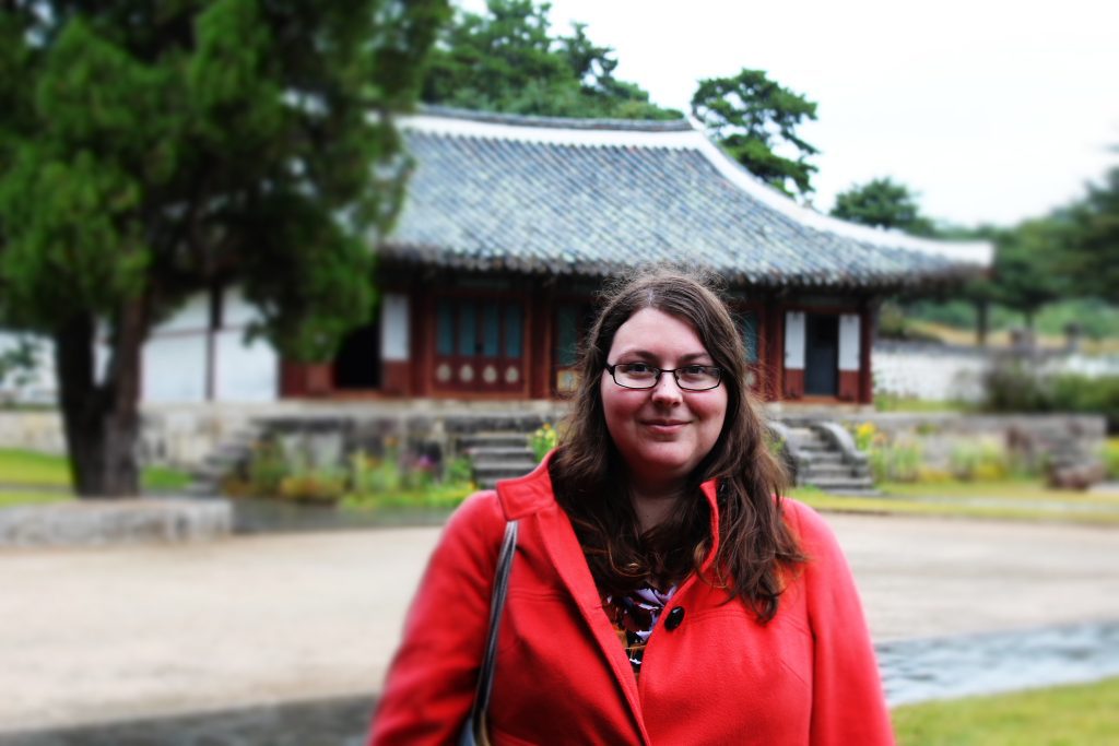 Photo of a woman in a bright red coat standing in front of a Korean building. The photo is cut off so you only see the upper part of her body. She has long brown hair and is wearing glasses.