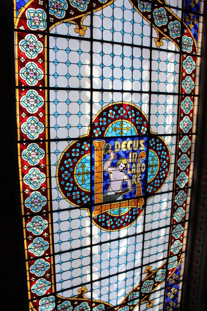 Photo of the stained glass ceiling. In the middle is a blacksmith and the words "Decus in Labore". There is a flower border around the outside.