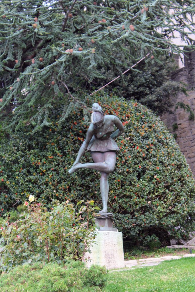 Bronze sculpture of a ballet dancer in a green, grassy area with lots of bushes.