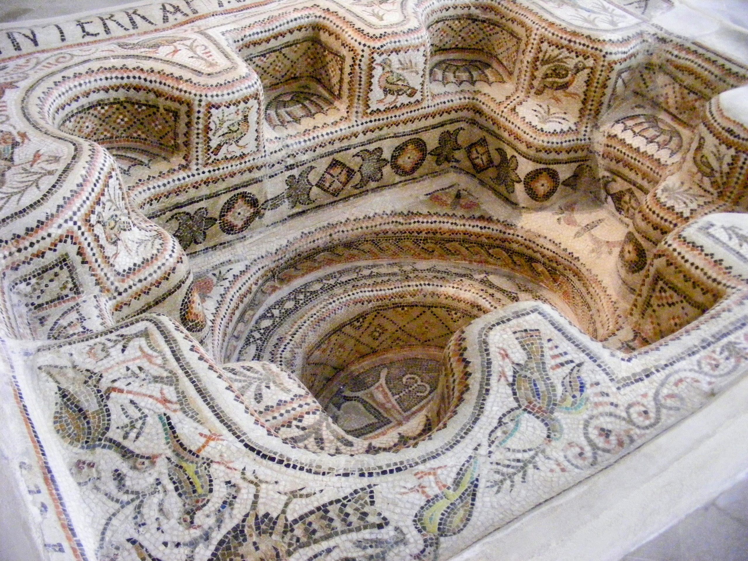 Photo of a 3D mosaic of a baptismal font. There is a deep hole in the middle with seats arranged around it.