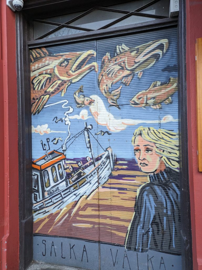 Photo of some street art in Reykjavik. The art shows a woman standing looking to the left. There is a boat in the background on the water with loads of giant fish swimming in the air.