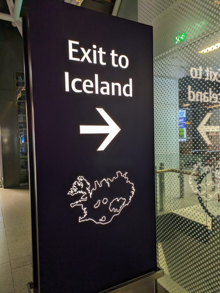 Photo of a sign that has an arrow pointing towards an escalator. The text reads "Exit to Iceland". Underneath the text is an outline of the island of Iceland.