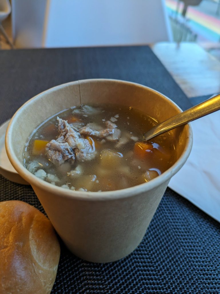 Photo of a takeaway cup of soup. You can see pieces of meat and carrot floating in a clear broth. There is a small bread roll at the bottom left and a spoon sticking out of the cup. Through the window, you can see the rainbow on the ground from Rainbow Street.