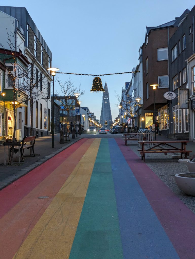 Photo of the Rainbow Street in Reykjavik. The street is painted with a rainbow leading to a church in the background. There are benches either side of the street and cars driving in the distance. The trees lining the street have fairy lights on them and there is a christmas decoration of a bell hanging above the street.