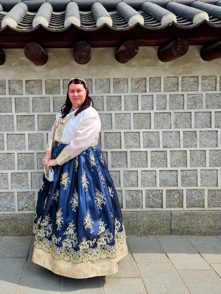 Photo of me standing in front of a brick wall wearing a Hanbok. The skirt is dark blue with golden patterns and I'm wearing a white jacket with matching gold patterns on the sleeves. I have my hair tied back and a silver hairband and am holding a matching purse.
