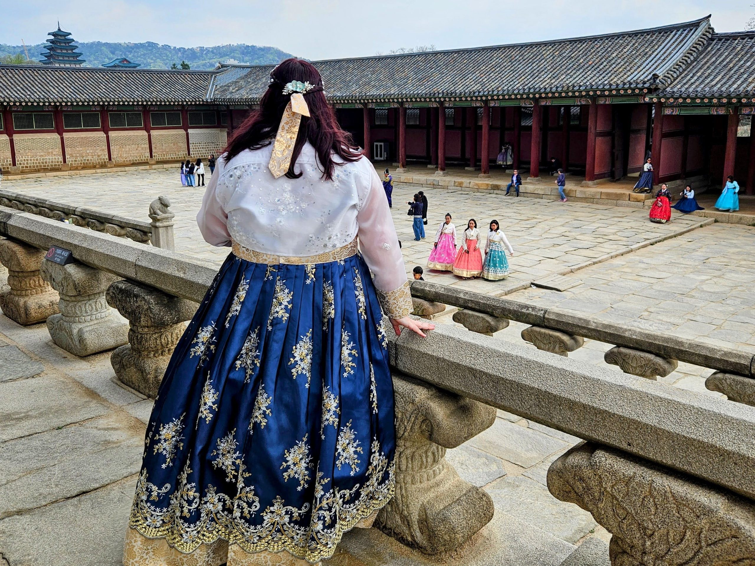 Photo of me wearing plus size Hanbok at Gyeongbokgung palace. I'm standing on a ledge and below are more people wearing hanbok posing for photos.