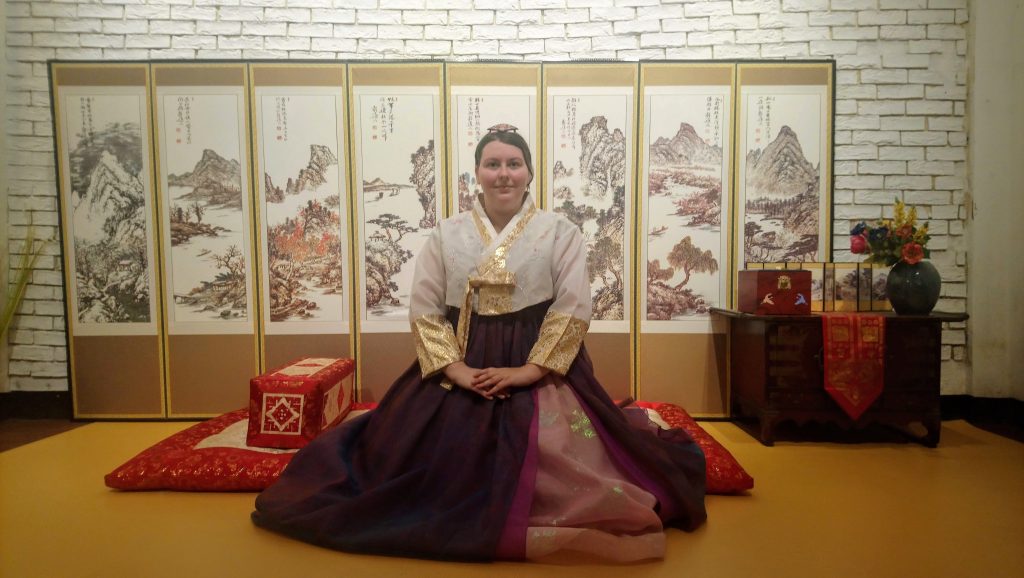 Photo of me wearing a purple hanbok sitting on the floor in front of a traditional Korean screen. The screen has eight panels showing mountains. I am sat with my hands resting on my lap and my hair is up in a traditional Korean style with a headpiece.