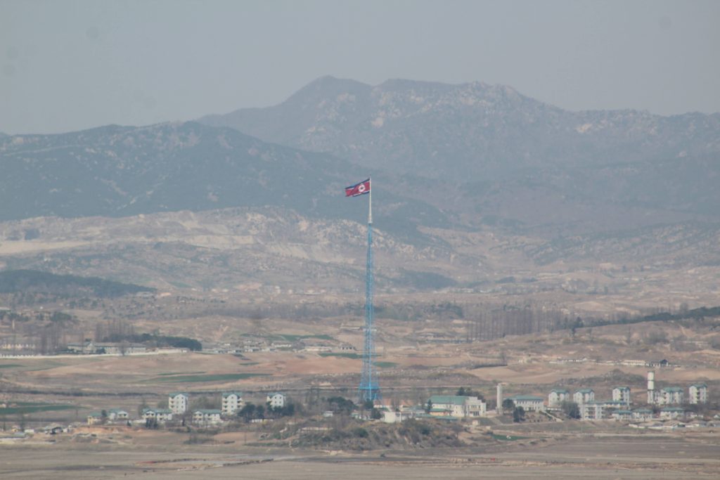 Photo of a small village. In the middle of the village is a very large flagpole flying the North Korean flag. In the background are distant mountains.