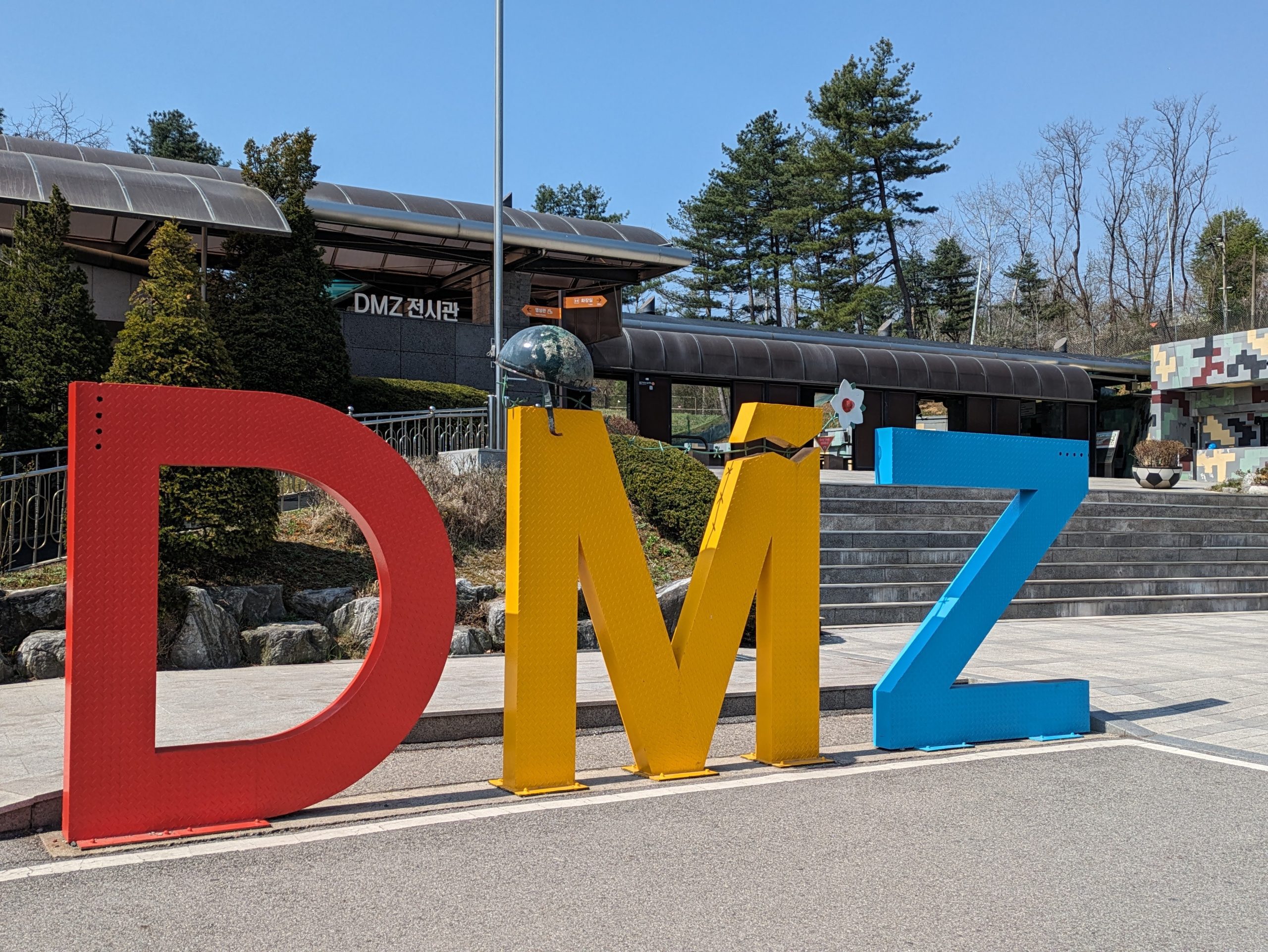 Photo of giant letters outside a building spelling out DMZ. The D is red, M is yellow and Z is blue. A helmet rests on the M and behind it is a flagpole.