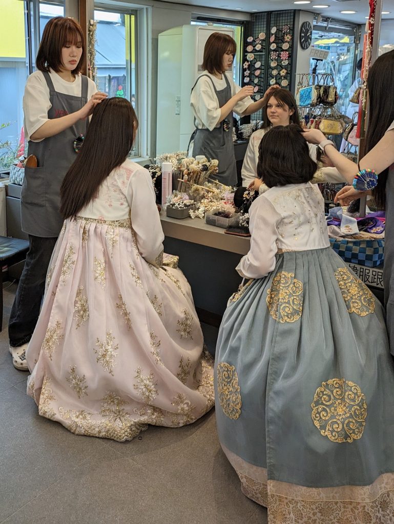 Photo shows two girls wearing Hanbok getting their hair done up. The girl on the left is wearing a light pink hanbok and has long brown hair that the hairdresser is about to touch. The girl on the right has shorter hair and the hairdresser is holding a section of it.