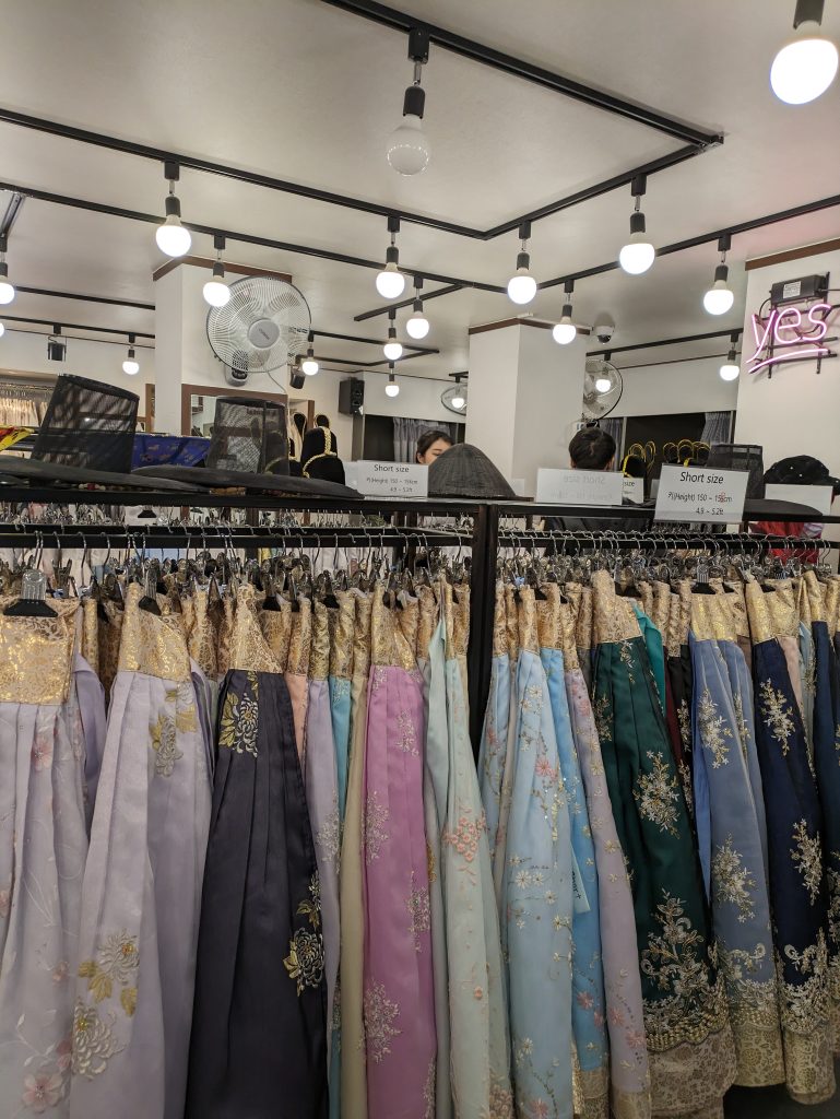 A photo of a rail in a shop with a selection of Hanbok in different colours. A sign above the rail says "short size". On the wall in the background is a sign saying "Yes". 