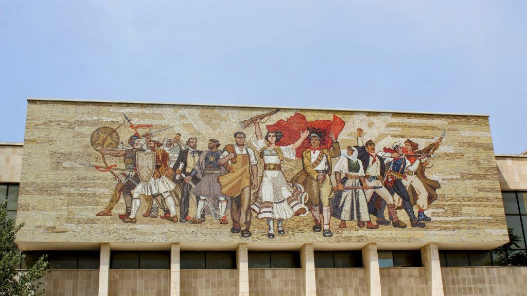 Photo of the outside of a building. There are various people in different historical outfits with one carrying the Albanian flag and several others with weapons such as guns and swords.
