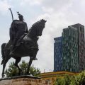 Photo of a man riding a horse. He is holding a sword in his right hand and there is a bird sitting on top of both the sword and on top of his hat. In the background are three tall rectangular buildings, one in blue, one in green and one in a dark orange colour.