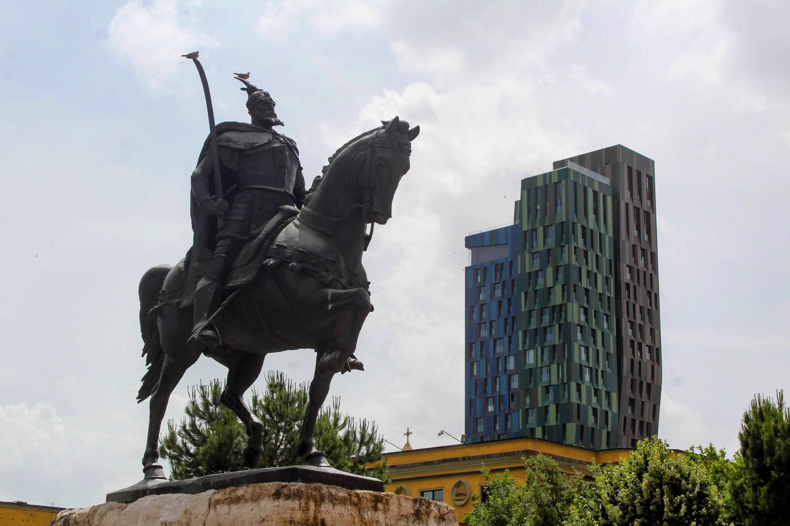 Photo of a man riding a horse. He is holding a sword in his right hand and there is a bird sitting on top of both the sword and on top of his hat. In the background are three tall rectangular buildings, one in blue, one in green and one in a dark orange colour.