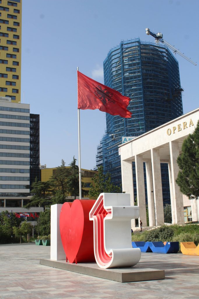 Photo of large letters saying "I heart t". Behind the letters is a large flagpole flying the Albania flag. To the right is a building with rectangular columns that says Opera and behind that is a building covered in scaffolding.