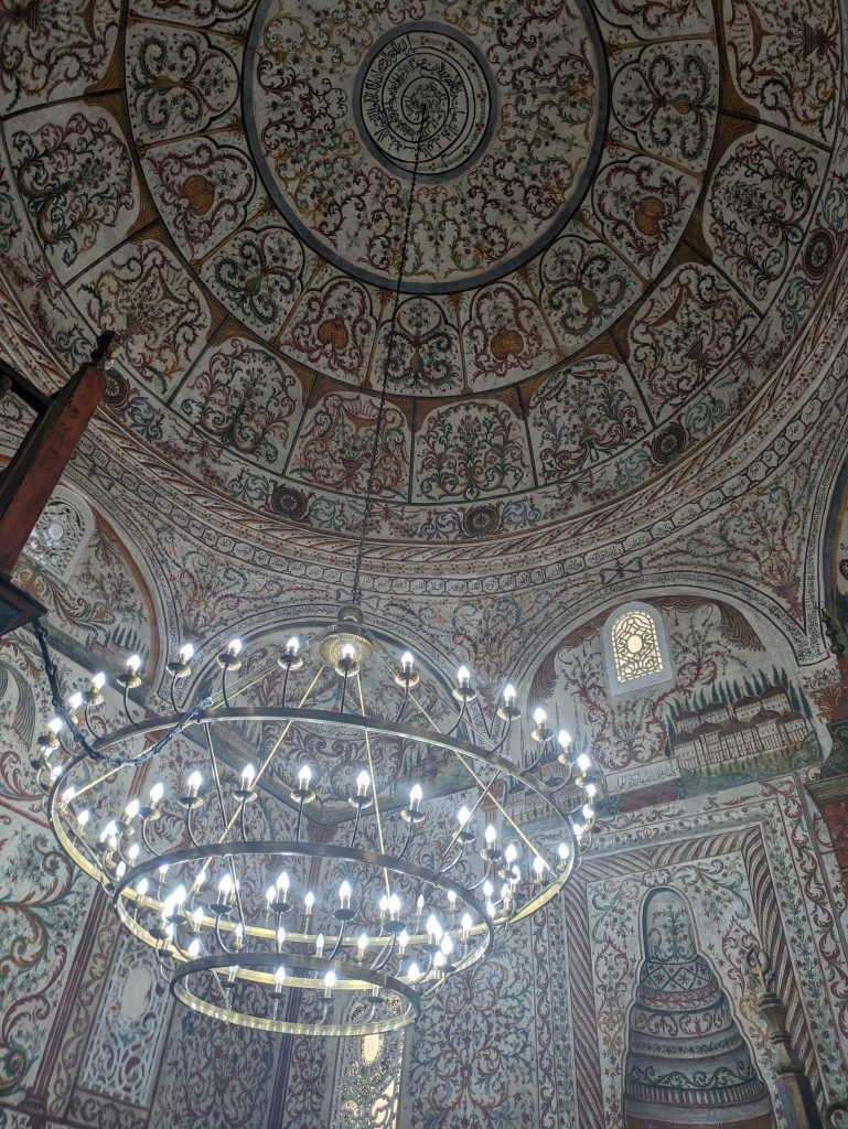 Photo of the inside of a mosque. The walls and ceiling are all decorated with paintings, lots of beautiful floral patterns and some buildings. There is also a large chandelier hanging from the roof.