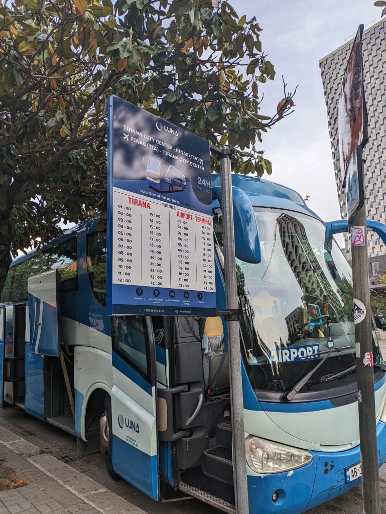 Photo of a blue bus with a sign in the window saying airport. In front of the bus is a timetable showing the airport times which are on the hour, every hour.