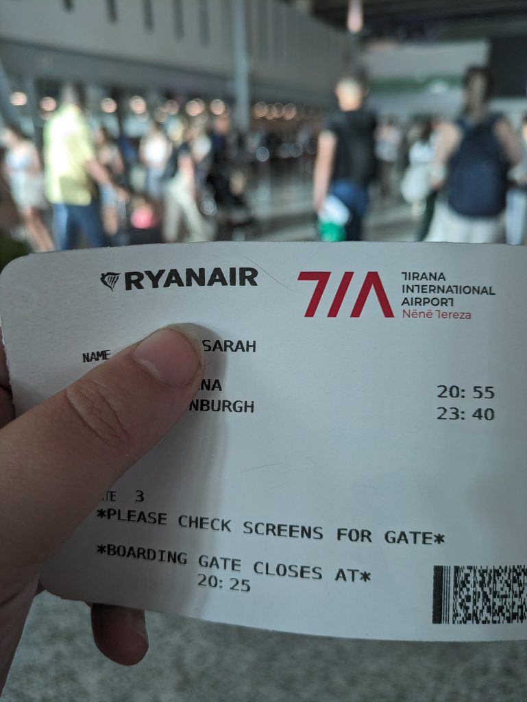 Photo of a Ryanair boarding pass. It says Tirana international airport and the name says "Sarah". It is being held so that you can't see the last name. The destination is partially covered so only the letters "nburgh" are visible.