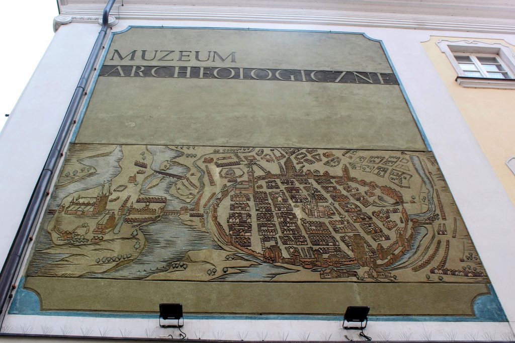 Photo of a wall that says Muzeum Archeologiczne. Underneath the words is a large map showing the old city of Poznan. There is an old town surrounded by circular walls with the town hall in the middle. On the left, outside the walls are two rivers with an island in the middle with a cathedral on it. There are some farms to the north of the city walls and another river to the east. In the top right of the photo is a small window to give some scale to the image. The window is around ten times smaller than the map but is a normal sized window.