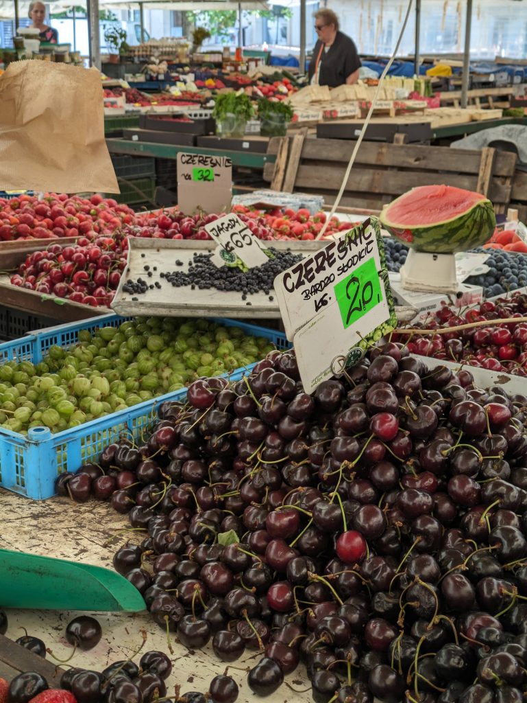 Photo of a market stall. There is a wide variety of different fruits on the table such as cherries, gooseberries, strawberries and blueberries. There are signs in Polish with what is likely the names of the fruit and prices showing how much it is for one kg of each item. In the background is another market stall with a woman walking between the stalls. 