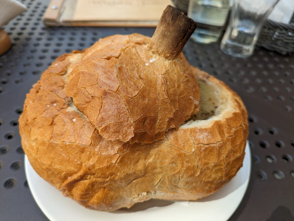 Photo of a loaf of bread that is in the shape of an apple. The top part is cut off and at a slight angle, letting you see the rest has been hollowed out and is filled with a clear soup. 