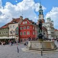 Photo of the old town square of Poznan. In the foreground is a fountain. Behind it is a row of colourful houses and the town hall in the background.