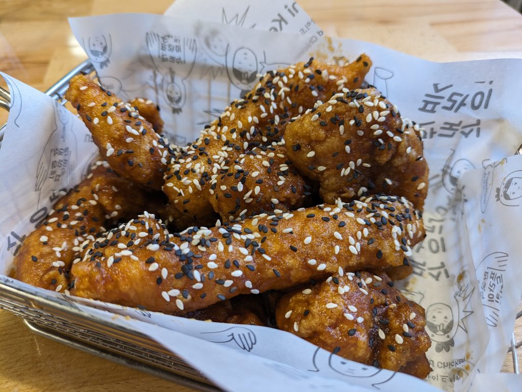 Photo of strips of chicken that have been deep fried. They are covered in black and white sesame seeds and in a box with wrapping showing korean characters and a person in a chicken outfit.