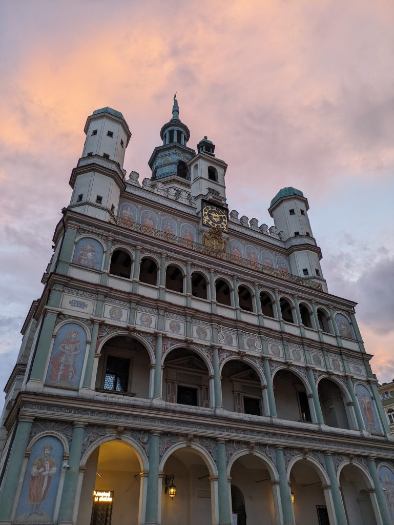 Photo of Poznan town hall with a soft glowing pink sky. The town hall has three levels of arches getting smaller as it goes up the building with lots of portraits of faces and full size historical people. At the top is a clock with gold hands and a small wooden door above the clock. Above that is a tower. There are also two smaller towers on either side of the building.