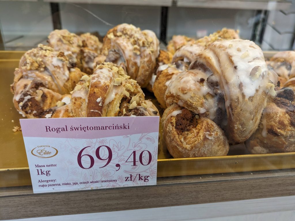 Photo of St Martin's croissant with a sign in front with the name in polish. It says 69,40 zloty per kg and lists the allergens in Polish. The logo for the bakery, Cukiernia Elite is also on the sign. The croissants are covered in what looks like white icing and nuts.