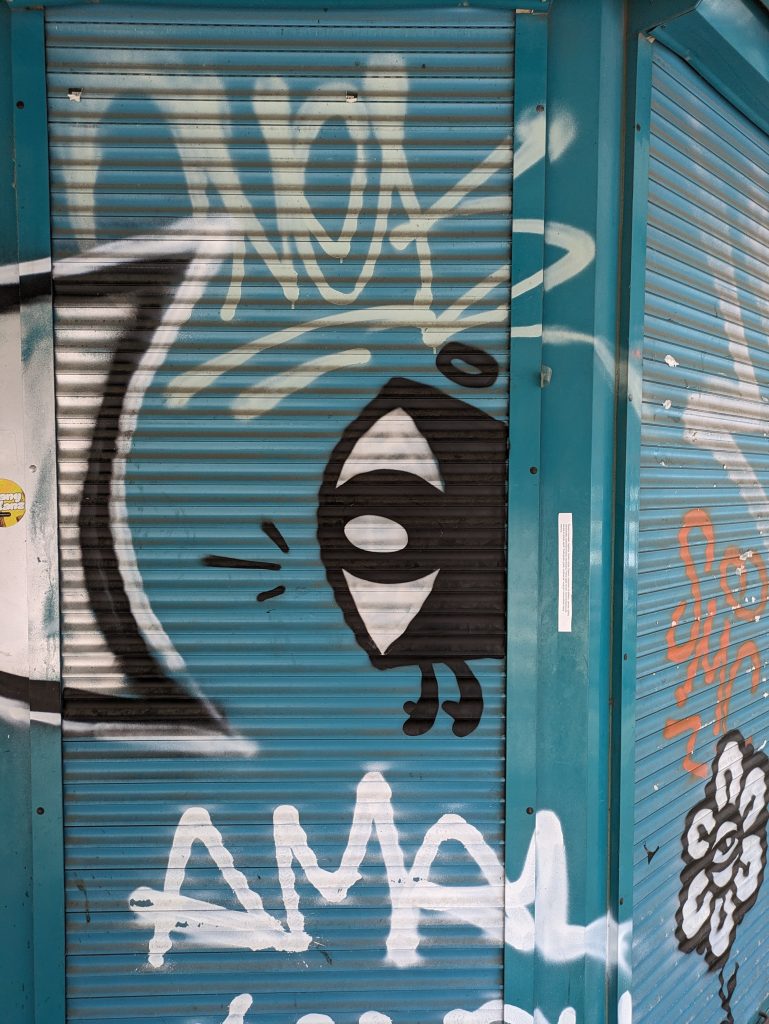 Photo of the same character with the eye for a face, now painted on a blue door with other graffiti surrounding it.