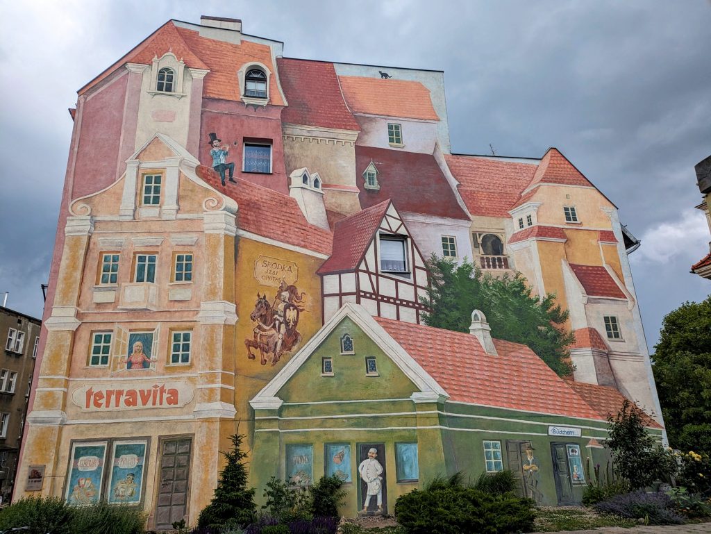 Photo of the side of a building which has been painted to look as if it's a collection of different buildings. At the front is a small green house, with a yellow house to the left and several others of varying sizes behind them. The mural covers almost the entire building, with only a few bits at the top and on the right that remind you its art.