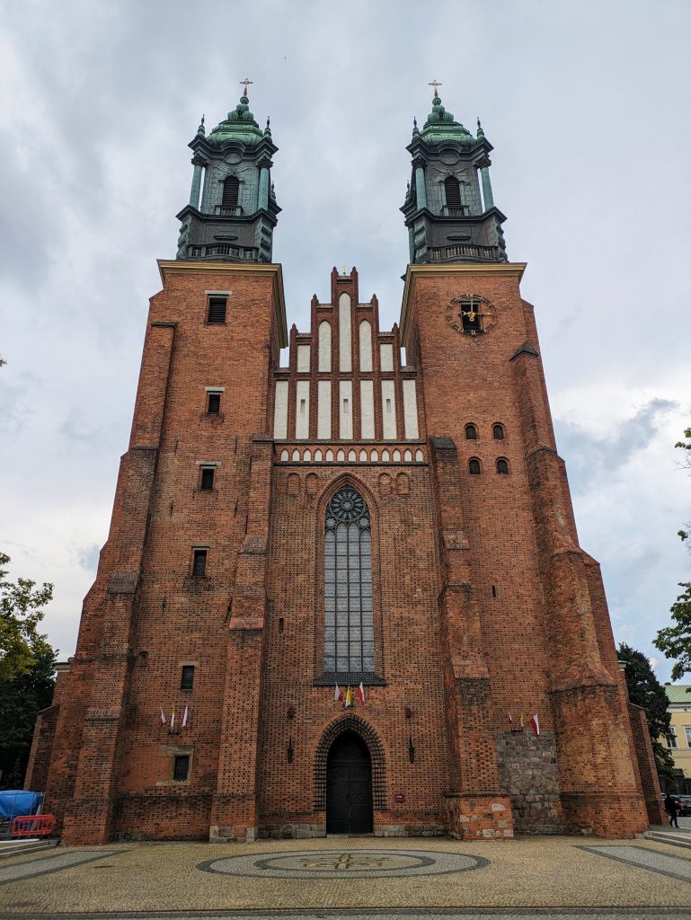 Photo of Poznan Cathedral. It is a very tall building with towers on both sides. The top of the tower switches from being red brick to being a greenish colour looking like it's made of bronze. There is a singular tall arch shaped window in the middle. On the left are some small windows regularly spaced up the column while the right coloumn only has a group of four small windows halfway up . Flags are hanging in front of the door and on either side, it's hard to make out what they are but one is clearly the Polish flag. 