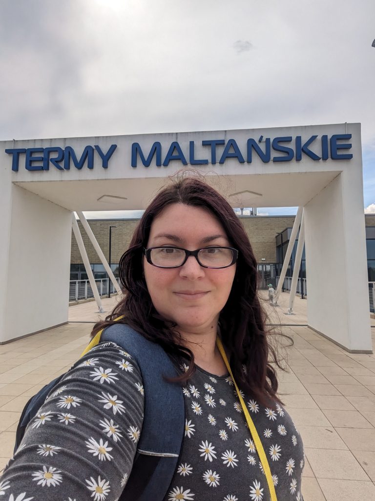 Selfie of me, wearing a black dress with daisies on it. I am standing in front of a rectangular arch that says Termy Maltanskie on a walkway leading to a long building in the background.