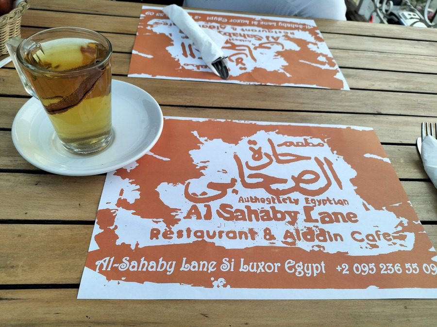 Photo of a table with a cup of what looks like tea with a cinnamon stick in it. On the table are two placemats that read "Authenticly Egyptian Al Sahaby Lane Restaurant and Aladdin Cafe". It then gives the address as Al-Sahaby Lane Si Luxor Egypt and the phone number which is +2 095 236 55 09