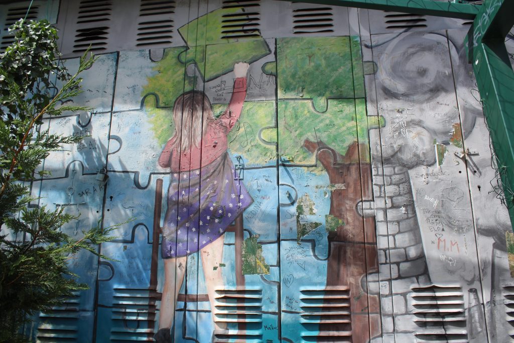 Photo of some street art. The art features a girl on a placing a jigsaw piece into the background. The piece she is holding is green and is completing a tree. Half of it is a tree and the other side of the trunk is a brick chimnew with smoke. THere are quite a few spots on the art where people have written things like their names as additional griffiti.