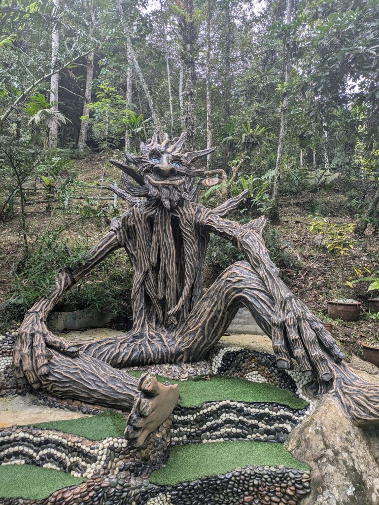 Photo of a large sculpture of what looks like a tree person. Behind the sculpture is forest, meanwhile the sculpture is sitting on an area with what looks like fake grass and steps decorated with stones. The stones are in different colours and are arranged in wave patterns.