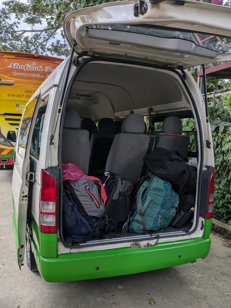 A photo of the Kota Kinabalu to Sandakan shuttle bus. The back door is open showing several backpacks stored in the small luggage area. Behind the shuttle is a much larger tour bus.