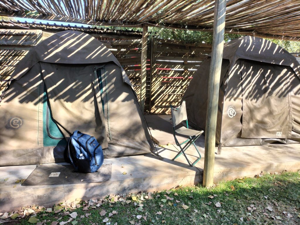 Photo of two tents underneath a wooden shelter. There is a wooden fence behind them and a backpack in front of one of the tents. There is a camping chair between the two tents. The tents are on a concrete surface, with a little bit of grass shown in the bottom right of the image.