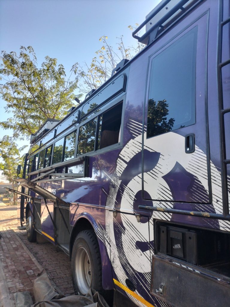 Photo of a large purple bus with several compartments at the side opened. On the ground are several tent bags. The bus has a large G in white on the side of it.