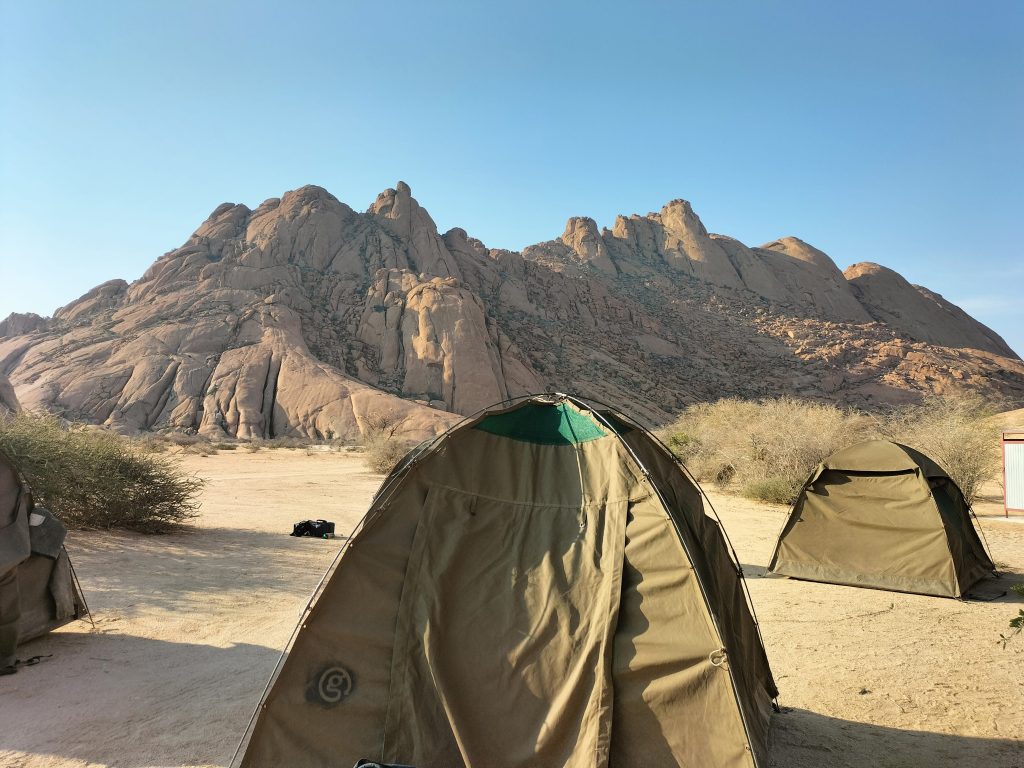 Photo of several tents sitting on a sandy ground. In the background is a large rocky outcrop that is quite high. The tents are facing different directions, but the one in the front faces the camera and has a g logo in the bottom left.