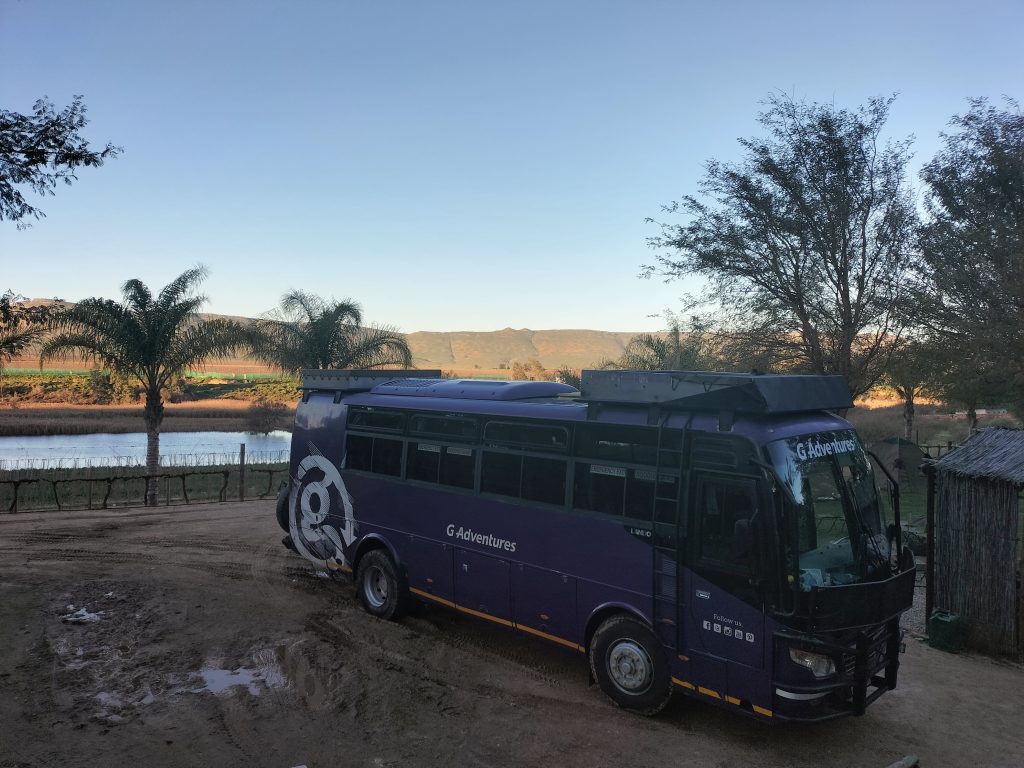 Photo of a large purple bus on a dirt road. In the background is some water with a few trees lining it. On the right, behind the bus, you can see a tent underneath some trees. There is a small shack between the bus and the tent. The bus itself has a large G in white on it, and it says G Adventures in the middle. There is a ladder leading up the side to the top to a storage container which is currently empty. This is the bus used on the Southern Africa tour with G Adventures.