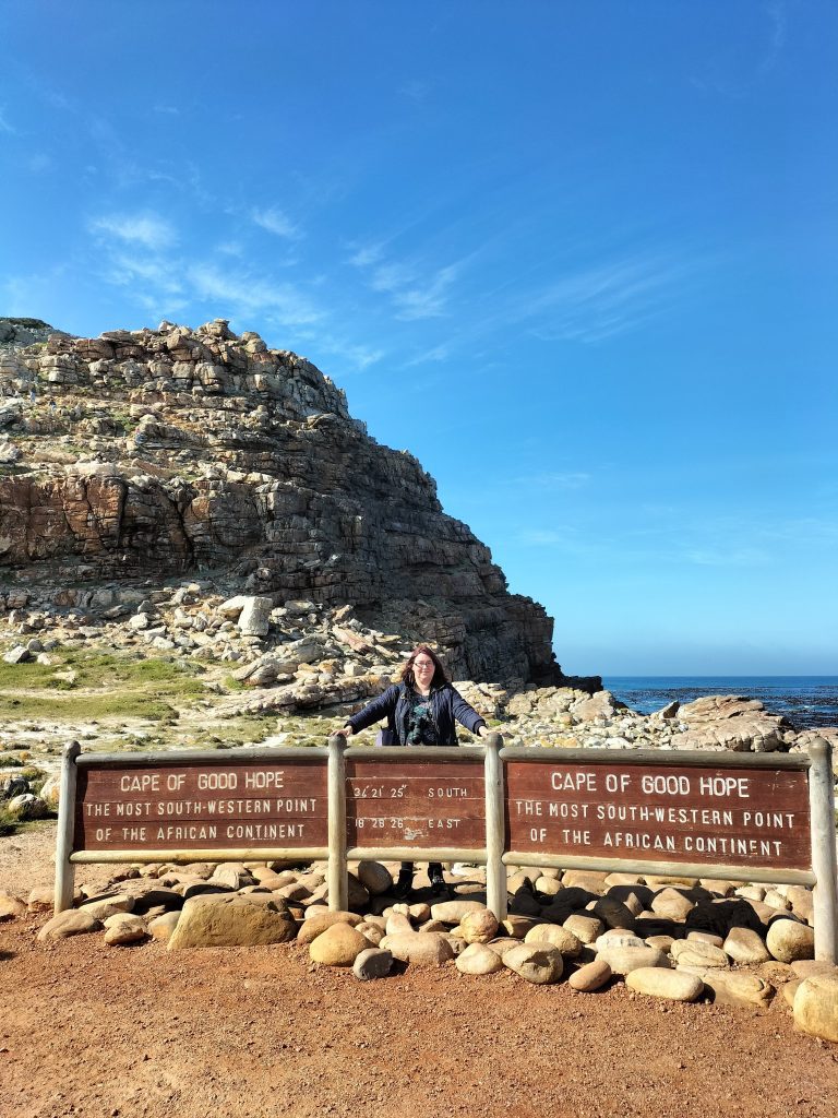 Photo of me standing behind a big wooden sign saying "Cape of Good Hope". Behind me are some rocks and to the right is the sea.