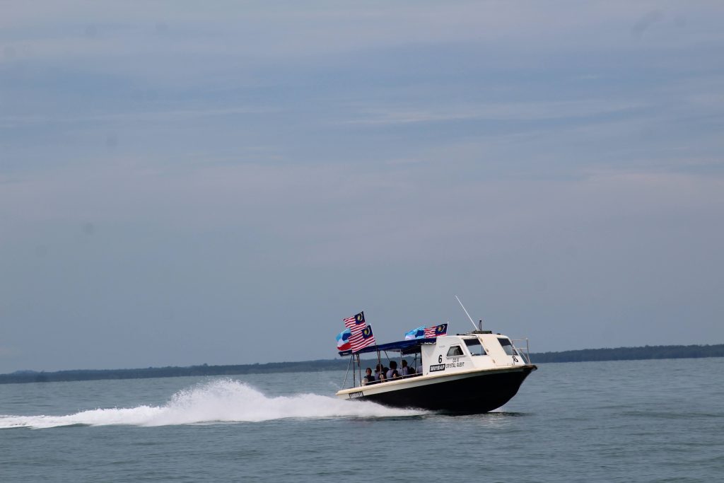 Photo of a speed boat in the sea heading towards Turtle Island. It has three malaysian flags flying from it, and two Sabah flags. The boat has the number 6 on it, and you can also see the words Crystal Quest.