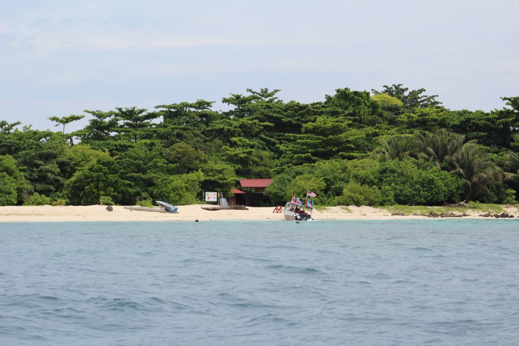 Photo of Turtle island from the water. There is one boat pulled up on the beach and another smaller boat pulled up further on the sand. Behind the beach is a lot of trees, very close together. There is a gateway with a red roof that leads into the forest.