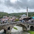 Photo of the iconic bridge in Prizren. Behind it you can see the mosque and some houses with lots of trees on the hills in the background.