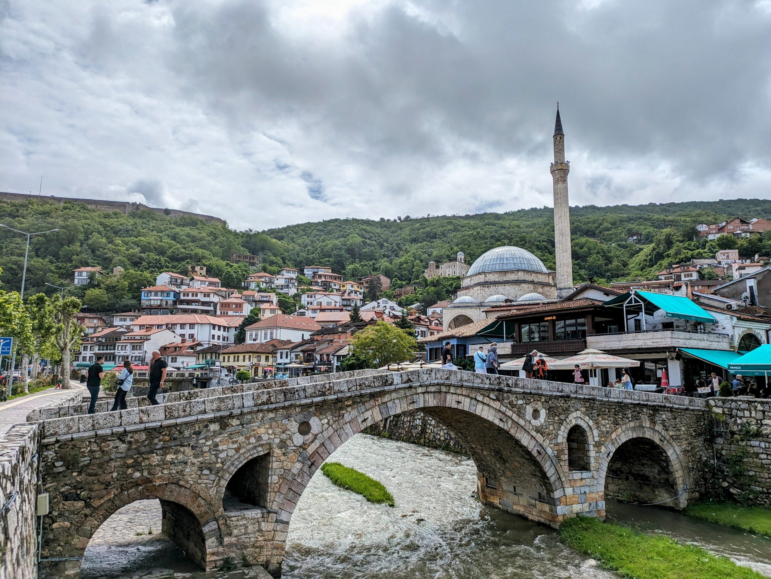 Photo of the iconic bridge in Prizren. Behind it you can see the mosque and some houses with lots of trees on the hills in the background.
