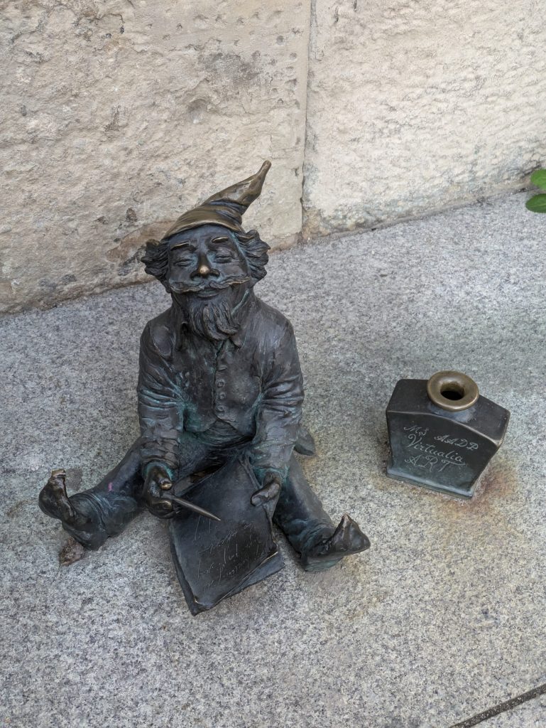 Photo of a dwarf statue. The dwarf is sitting on the ground, writing a letter. On the right is an ink bottle that says Virtualia Art.