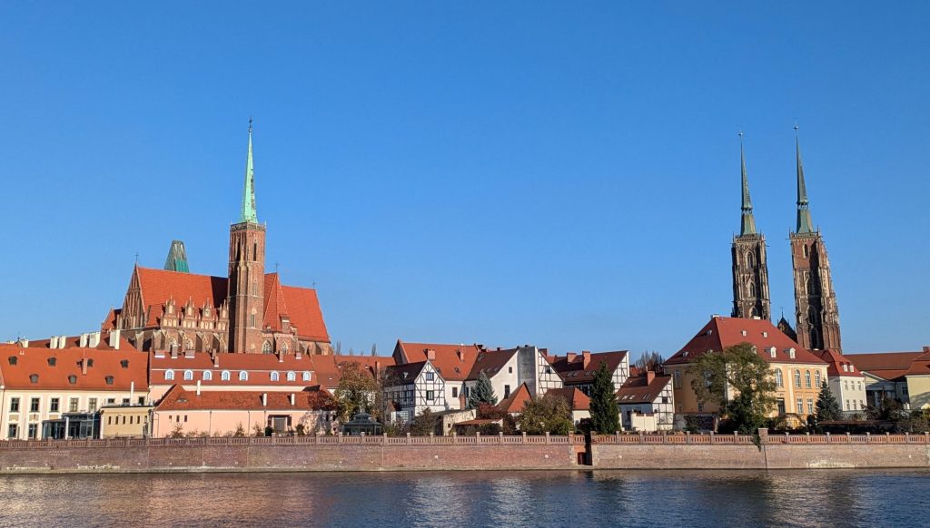 Photo looking across water to the skyline of cathedral island in Wroclaw. You can see two spires from a cathedral on the right, and another church on the left.