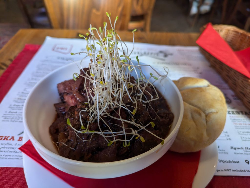 Photo of a bowl containing a meat stew. On the top is watercress. Underneath the bowl is a menu but it's too blurry to read anything, and a small bread roll is next to the bowl.
