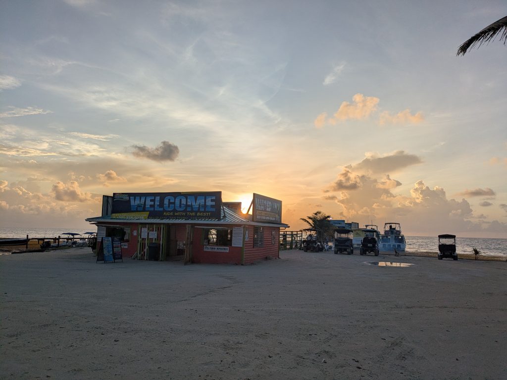 Photo of a small building with a big sign on the top that says "Welcome. Ride with the best". Below are two windows. Behind it is a pier with boats in the distance. You can make out one person behind the building, but aside from that it is completely empty. Behind the building you can see the sun has only just started to rise.